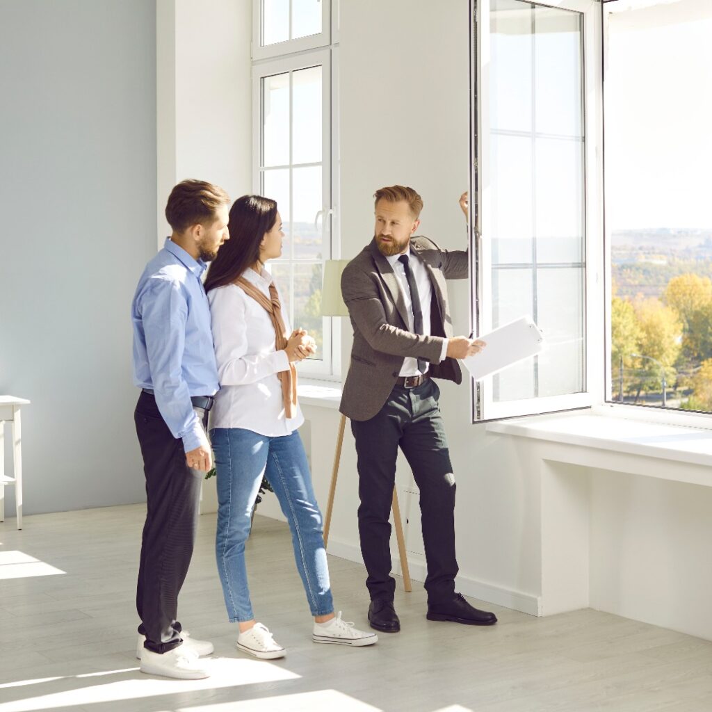 Handsome bearded male realtor in suit with documents in his hands advises young couple before buying new house, demonstrates view from the window. Three people are standing in modern sunny apartment.